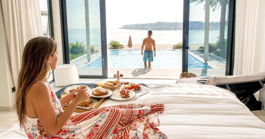 A lady enjoying the ultimate luxury of having breakfast in bed at a private villa with a pool overlooking the beach and sea as her partner takes a dip in the pool on a sunny day in Abu Dhabi