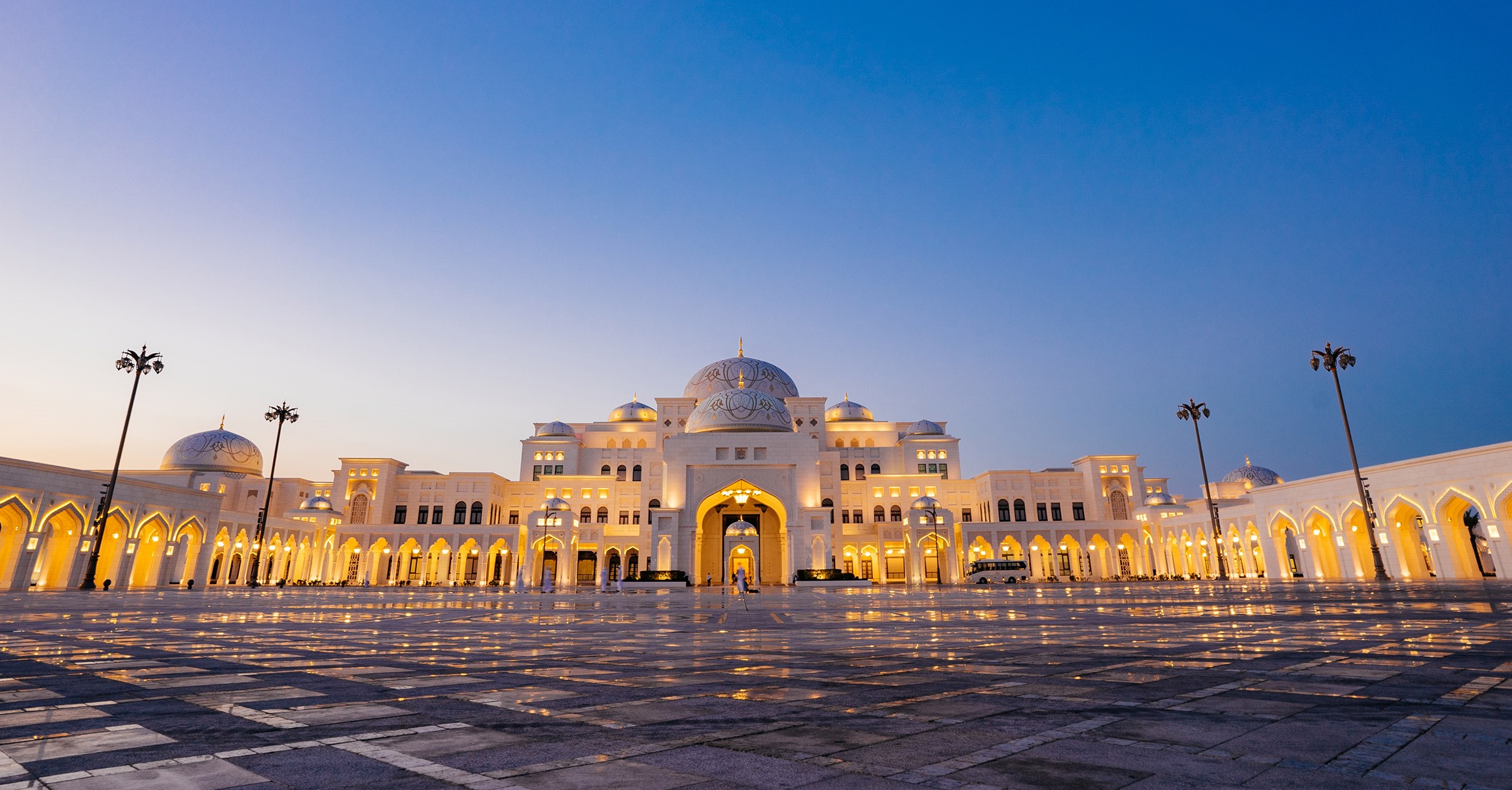 An exterior landscape shot of Qasr Al Watan Palace during sunset as the Palace is illuminated by bright yellow lights.