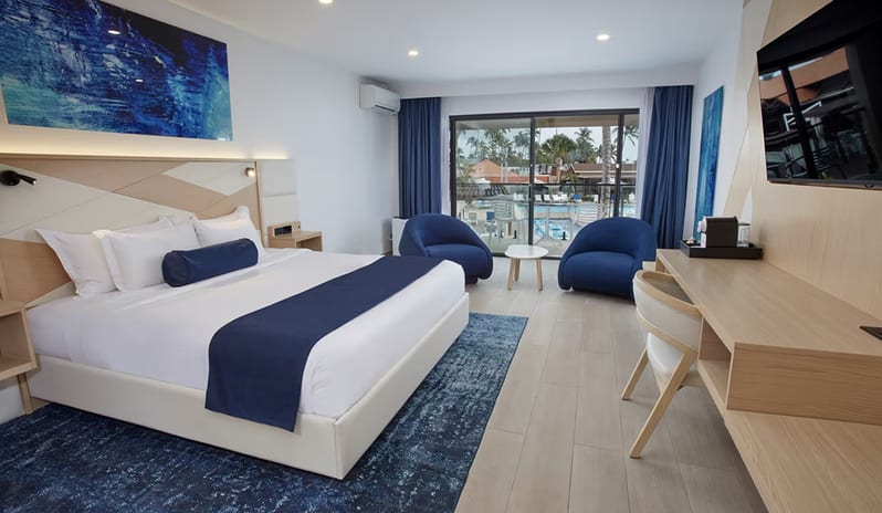 Pool View Room King bed, blue accents, plush armchairs, and a private balcony overlooking a tropical resort pool at Divi Aruba All Inclusive Resort by Kenwood Travel