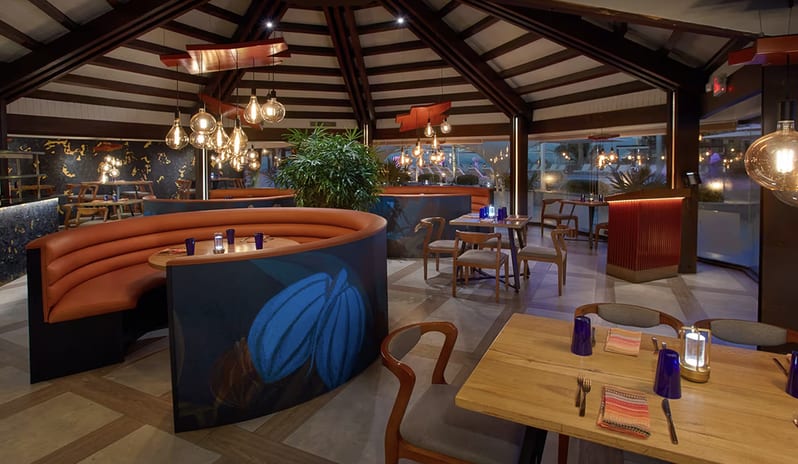 Pure Lime Sandals Royal Barbados featuring a high wood-beamed ceiling, modern Edison bulb light fixtures, and blue curved booths with orange leather seating at Divi Aruba All Inclusive Resort by Kenwood Travel