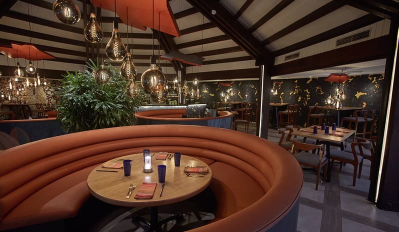 Pure Lime Sandals Royal Barbados featuring a high wood-beamed ceiling, modern Edison bulb light fixtures, and blue curved booths with orange leather seating at Divi Aruba All Inclusive Resort by Kenwood Travel