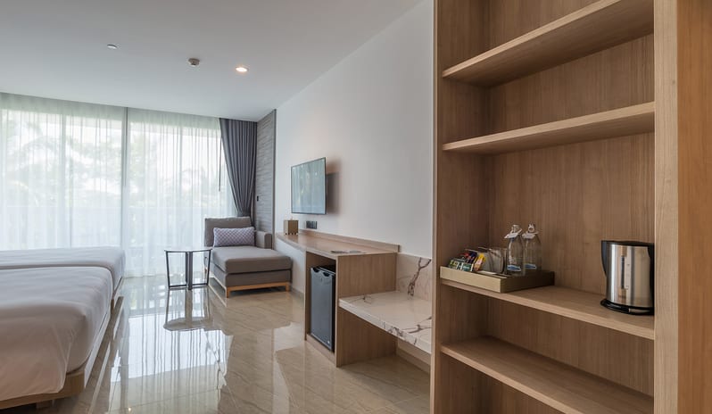 Deluxe Room interior showing light wood shelving with a kettle and coffee station, a wall-mounted TV, a small refrigerator, and a gray chaise lounge by a floor-to-ceiling window at Centara Ao Nang Beach Resort & Spa Krabi by Kenwood Travel