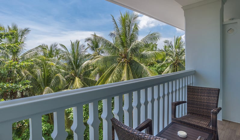Deluxe Roombalcony with woven wicker chairs and a small table, overlooking a lush garden of tropical palm trees under a bright blue sky at Centara Ao Nang Beach Resort & Spa Krabi by Kenwood Travel