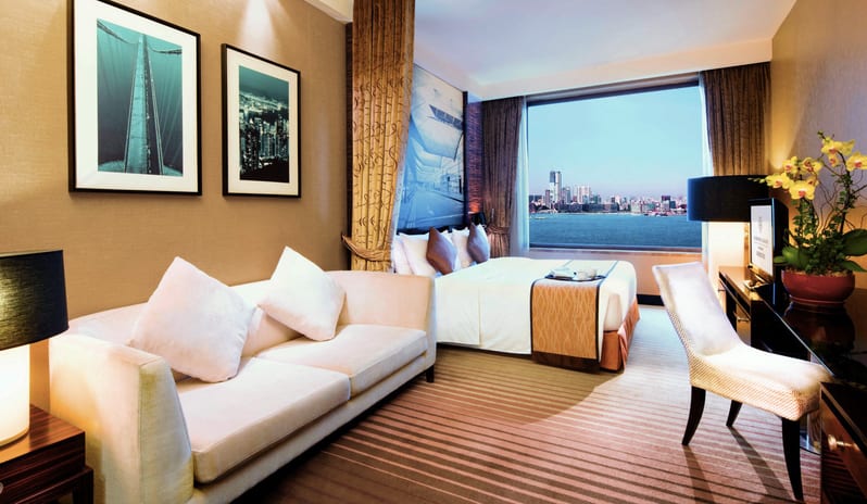 Premier Harbourview Club Room modern sofa, king bed, and a large window overlooking Victoria Harbour and the city skyline at Harbour Grand Hong Kong by Kenwood Travel