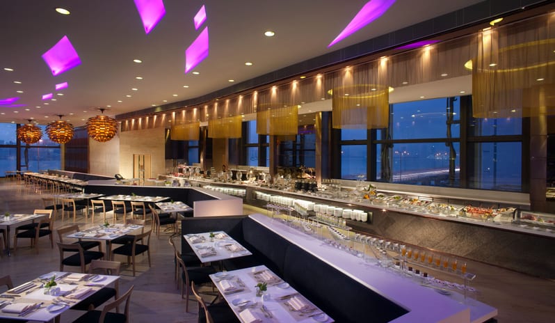 Harbour Grand Café long seafood buffet counter, purple geometric ceiling lights, and floor-to-ceiling windows overlooking Victoria Harbour at twilight at Harbour Grand Hong Kong by Kenwood Travel
