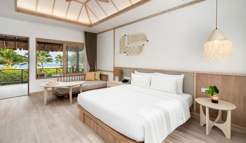 Deluxe Sea View Villa Bedroom featuring a white king-size bed, vaulted wood-beam ceiling, and large windows overlooking a beach and palm trees at SAii Phi Phi Island Village by Kenwood Travel