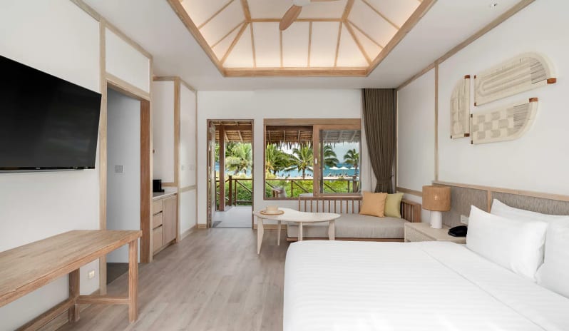 Deluxe Sea View Villa Bedroom featuring a white king-size bed, vaulted wood-beam ceiling, and large windows overlooking a beach and palm trees at SAii Phi Phi Island Village by Kenwood Travel