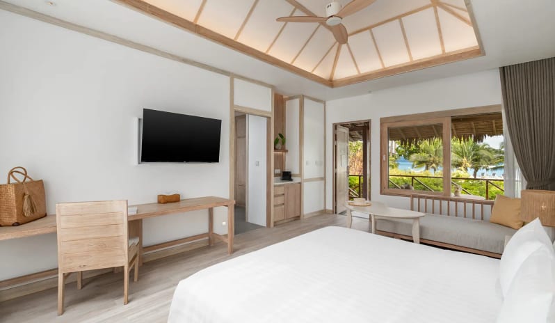 Deluxe Sea View Villa Bedroom featuring a white king-size bed, vaulted wood-beam ceiling, and large windows overlooking a beach and palm trees at SAii Phi Phi Island Village by Kenwood Travel