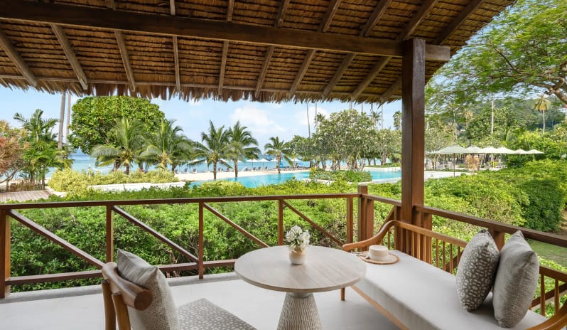 Deluxe Sea View Villa Daytime view from a private villa balcony with wooden furniture and a thatched roof, overlooking a lush tropical resort pool, white sand beach, and palm trees at SAii Phi Phi Island Village by Kenwood Travel