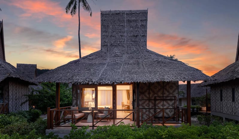 Deluxe Sea View Villa Exterior view of a luxury thatched-roof beach villa at sunset with warm interior lighting, a private wooden terrace, and a silhouette of a palm tree against a pink and orange sky at SAii Phi Phi Island Village by Kenwood Travel