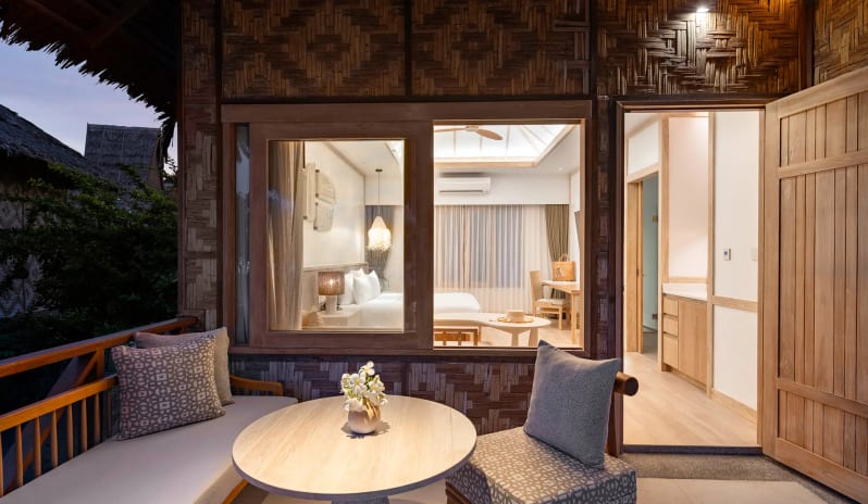 Deluxe Sea View Villa Close-up of a private villa terrace featuring a wooden table, cushioned chairs, and patterned pillows, with a view through the window into a warm at SAii Phi Phi Island Village by Kenwood Travel