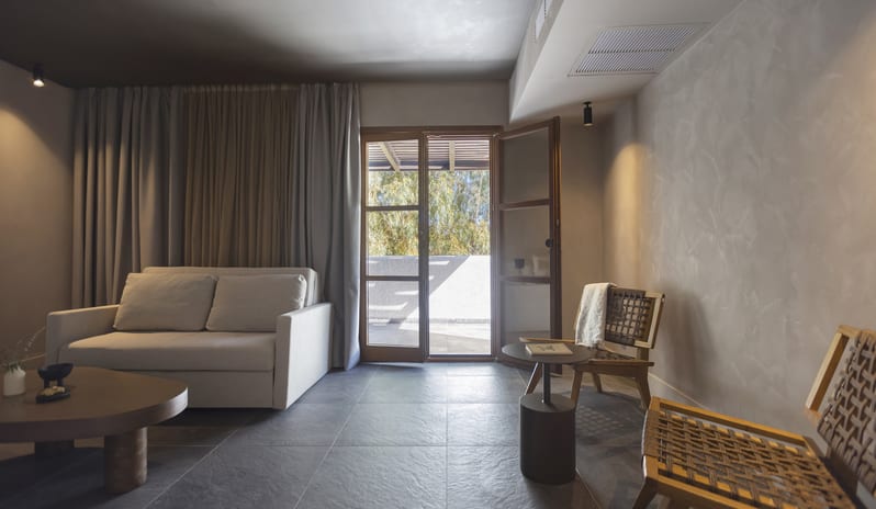 Tropical Bungalow Suite Spacious living area in a tropical bungalow suite featuring a cream sofa, organic-shaped wooden coffee table, and two woven accent chairs under warm spotlighting at Aulūs Lindos Rhodes, Curio Collection by Hilton by Kenwood Travel