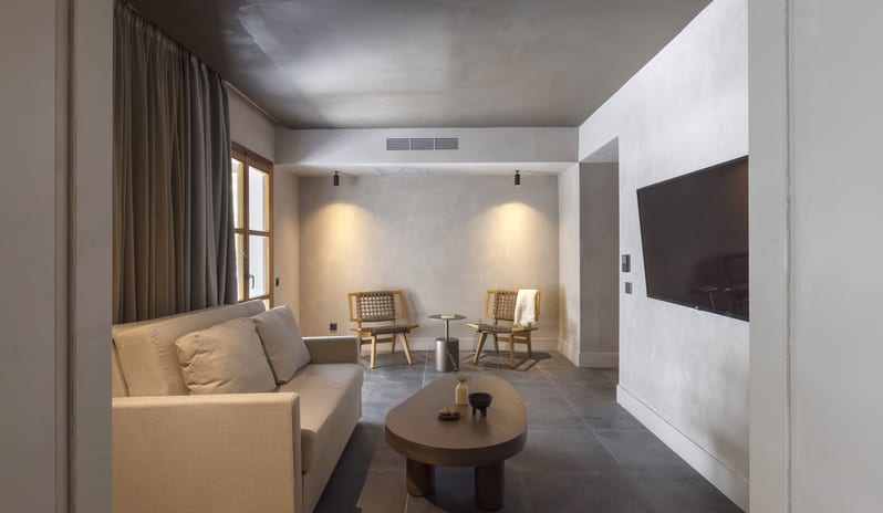 Tropical Bungalow Suite Spacious living area in a tropical bungalow suite featuring a cream sofa, organic-shaped wooden coffee table, and two woven accent chairs under warm spotlighting at Aulūs Lindos Rhodes, Curio Collection by Hilton by Kenwood Travel