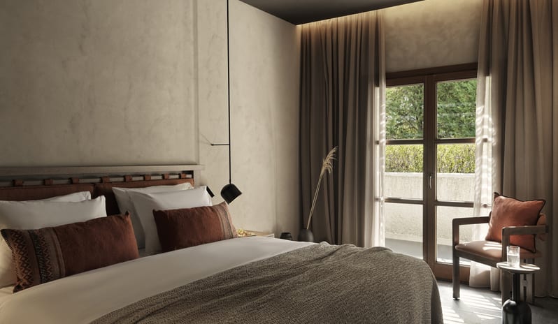 Hilton Tropical Retreat King Bed with Earthy linens, a Designer Accent Chair, and Natural Light from Large French Doors at Aulūs Lindos Rhodes, Curio Collection by Hilton by Kenwood Travel