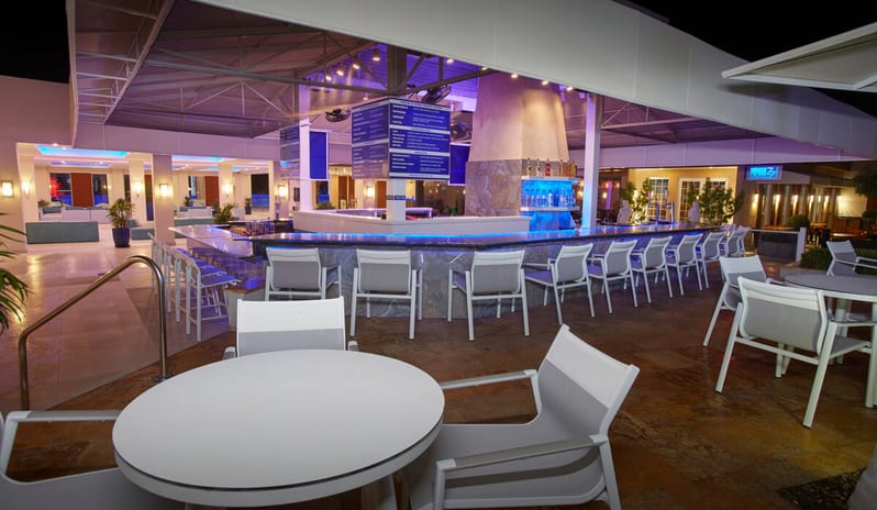 Pizza per Tutti outdoor beach bar at a luxury resort at night, featuring a sleek square counter with blue LED accent lighting, white bar stools, and palm trees under a dark sky in the background at Divi Aruba All Inclusive Resort by Kenwood Travel