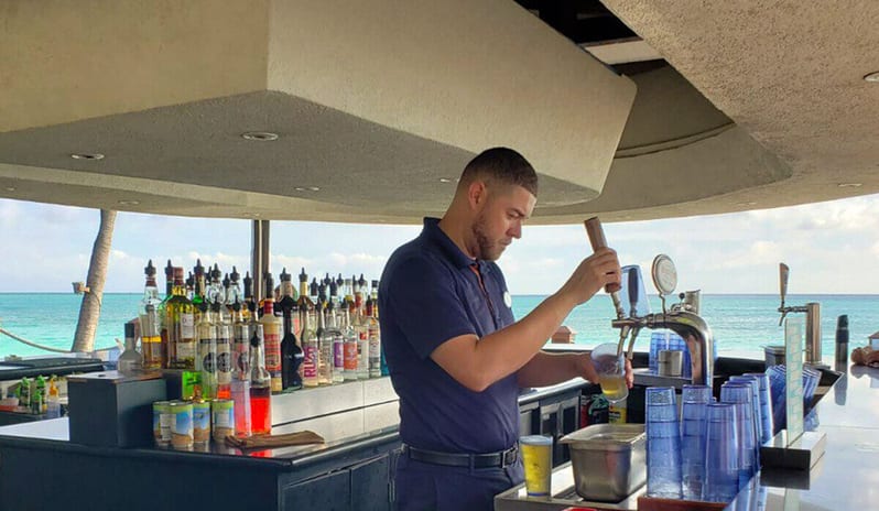 Pelican Bar A wooden beachfront deck at a Caribbean resort featuring white modern chairs, high-top tables, and rustic wood-slice tables at Divi Aruba All Inclusive Resort by Kenwood Travel