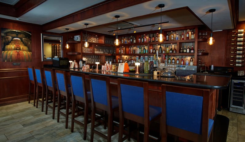 Paparazzi Restaurant, Bar & Lounge Interior view of a sophisticated cocktail bar featuring dark wood paneling, a black granite countertop, and a row of high-back blue upholstered bar stools under warm Edison bulb pendant lights at Divi Aruba All Inclusive Resort by Kenwood Travel