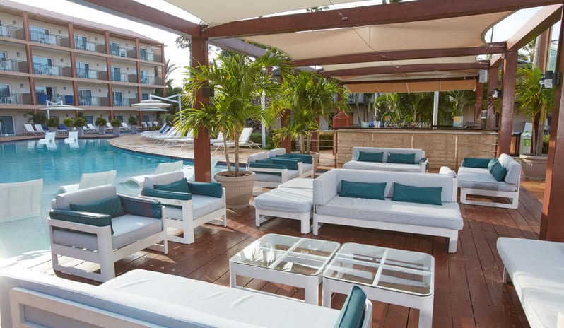 Poolside Bar patio seating area with white furniture and palm trees overlooking a blue resort pool at Divi Aruba All Inclusive Resort by Kenwood Travel