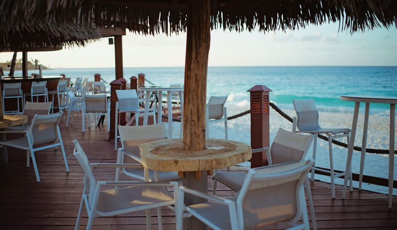 Pelican Bar outdoor beachfront bar and patio area with white modern chairs, wooden tables under thatched palapa roofs, and a view of turquoise ocean waves on a white sand beach at sunset at Divi Aruba All Inclusive Resort by Kenwood Travel