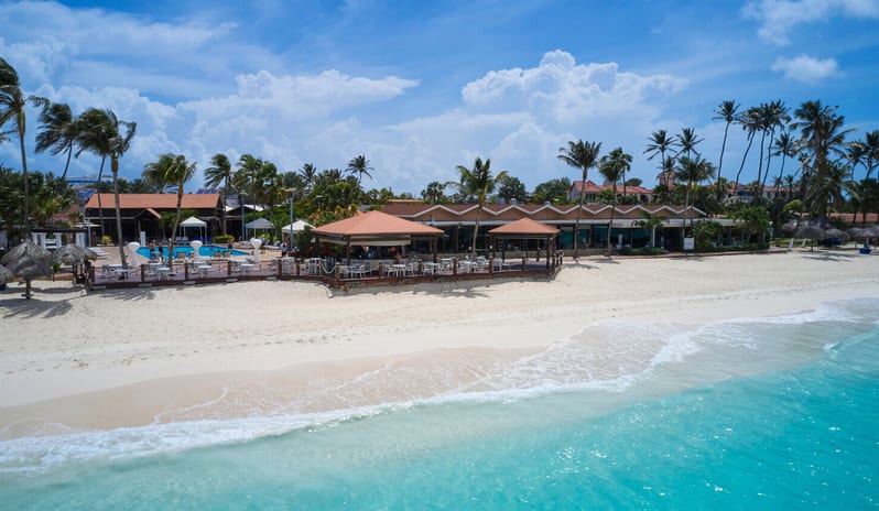 Pelican Bar luxury Caribbean beach resort featuring a turquoise ocean, white sandy beach, outdoor poolside bar with orange roofs, and palm trees under a blue sky at Divi Aruba All Inclusive Resort by Kenwood Travel