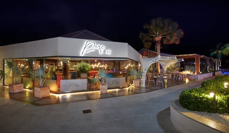 Pure Lime restaurant at night, featuring modern glass architecture, warm hanging lights, and outdoor patio seating under palm trees at Divi Aruba All Inclusive Resort by Kenwood Travel