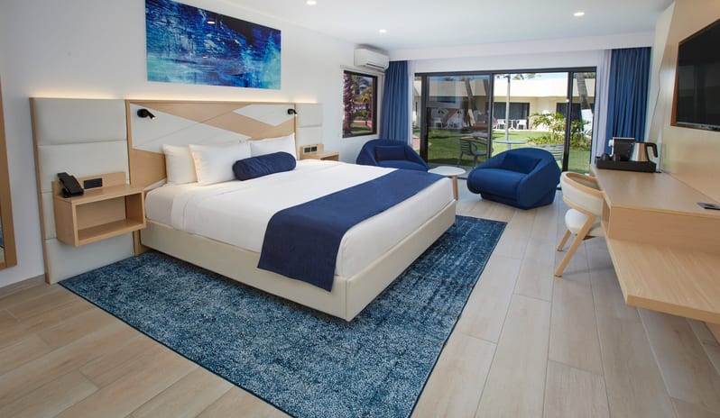 Beachside Room King bed, blue abstract wall art, and a large sliding glass door opening to a patio and garden view at Divi Aruba All Inclusive Resort by Kenwood Travel