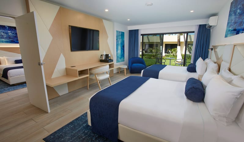 Beachside Room Queen Queen Spacious adjoining hotel rooms featuring a double queen bedroom in the foreground and a king bedroom visible through an open door, both with modern blue decor and garden views at Divi Aruba All Inclusive Resort by Kenwood Travel