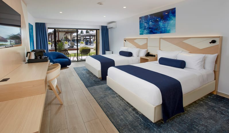Pool View Room Queen Queen room with two queen-sized beds, blue bed runners, and a private balcony offering a view of palm trees and the ocean at Divi Aruba All Inclusive Resort by Kenwood Travel