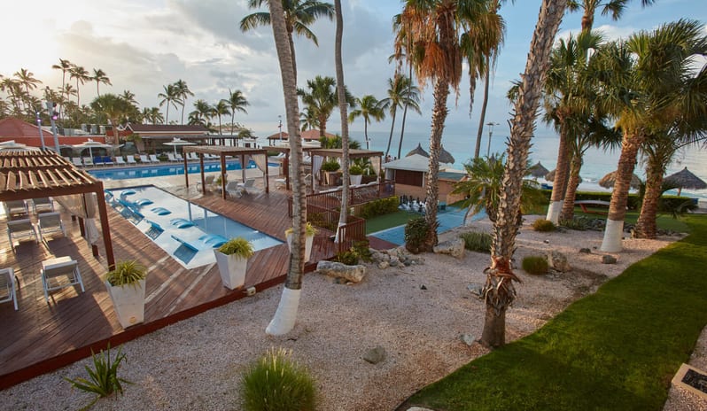Pool View Room Queen Queen room Aerial view of a luxury tropical resort featuring an oceanfront pool deck with wooden cabanas, palm trees, and lounge chairs near the beach at sunset at Divi Aruba All Inclusive Resort by Kenwood Travel