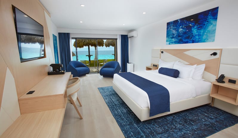 Ocean View Room King bed, blue accents, and a sliding glass door opening to a turquoise ocean view with palm trees at Divi Aruba All Inclusive Resort by Kenwood Travel