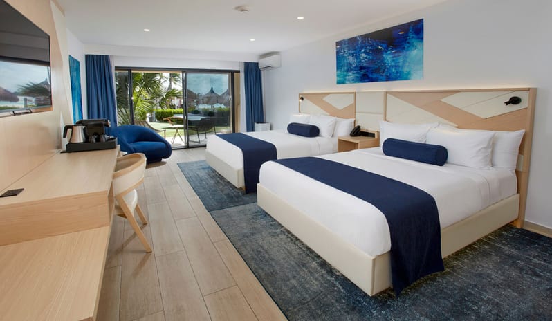 Ocean View Room Queen Queen beds, blue runners, a light wood desk, and sliding glass doors leading to a tropical patio and beach view at Divi Aruba All Inclusive Resort by Kenwood Travel