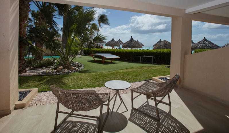 Ocean View Room Queen Queen patio with two wicker chairs and a small table looking out onto a grassy garden area, palm trees, and the ocean beach with straw umbrellas at Divi Aruba All Inclusive Resort by Kenwood Travel