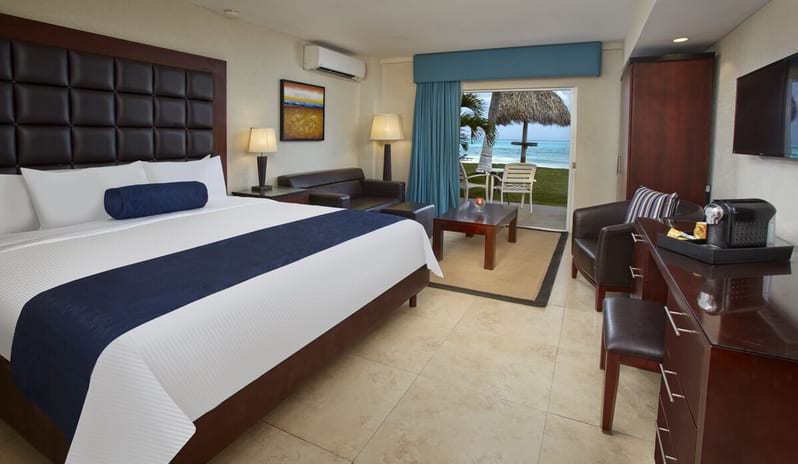 Oceanfront Lanai Room King bed with navy accents, a seating area, and sliding glass doors opening to a private terrace with an ocean view at Divi Aruba All Inclusive Resort by Kenwood Travel