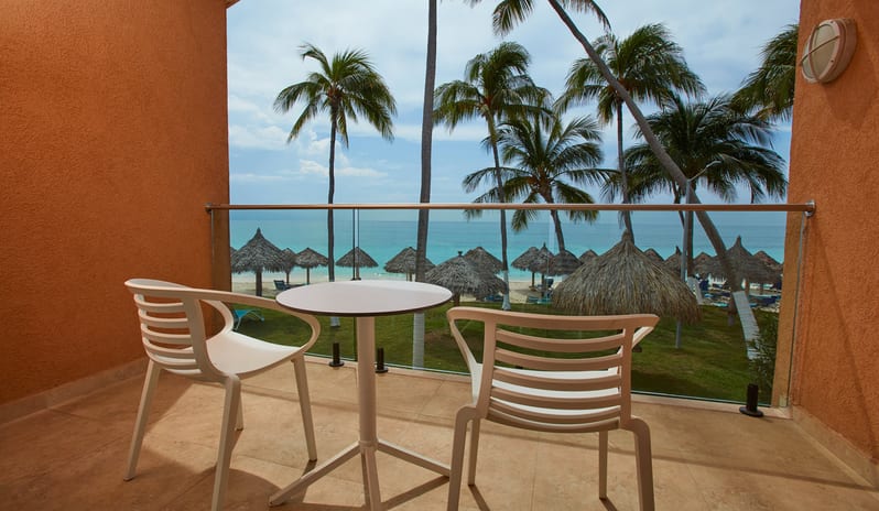 Oceanfront Lanai Room Queen Queen balcony with two white chairs and a small table, overlooking a tropical beach with turquoise water, palm trees, and thatched umbrellas at Divi Aruba All Inclusive Resort by Kenwood Travel