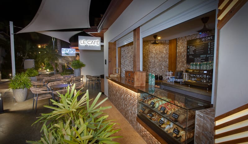 Le Caféat night, featuring a lit pastry display case filled with croissants, an espresso machine, and a patio with wicker chairs under a white sail canopy at Divi Aruba All Inclusive Resort by Kenwood Travel
