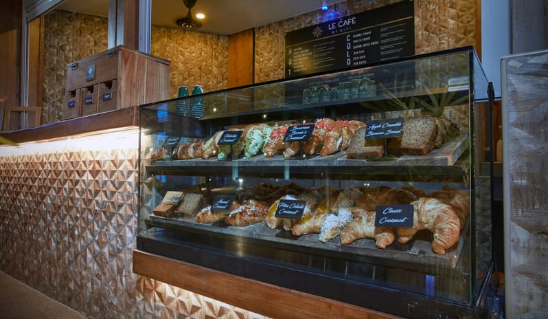 Le Caféat various gourmet pastries, including pistachio croissants, Bavarian berry croissants, apple crumble banana bread, and classic croissants, set against a textured wood-panel counter at Divi Aruba All Inclusive Resort by Kenwood Travel