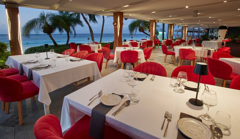 Red Parrot restaurant interior featuring red velvet chairs, white tablecloths, spiky black chandeliers, a blue-lit wine cellar wall, and a vertical green living wall at Divi Aruba All Inclusive Resort by Kenwood Travel