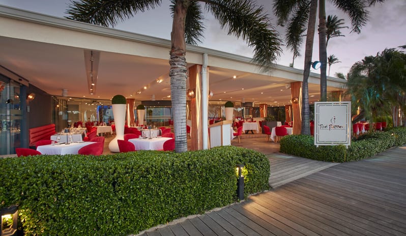 Red Parrot Outdoor seating at the Red Parrot restaurant featuring white tablecloths, red chairs, and palm trees along a wooden boardwalk at sunset at Divi Aruba All Inclusive Resort by Kenwood Travel