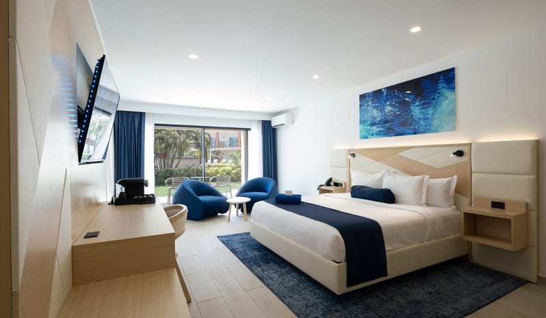 Garden View Room King bed with navy blue accents, a minimalist wooden desk, and two blue lounge chairs facing a private outdoor terrace at Divi Aruba All Inclusive Resort by Kenwood Travel