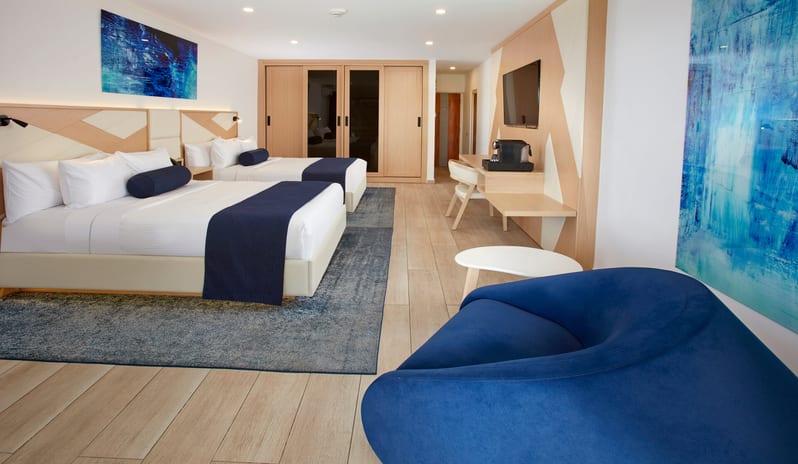 Pool View Room Queen Queen room with two queen-sized beds, blue bed runners, and a private balcony offering a view of palm trees and the ocean at Divi Aruba All Inclusive Resort by Kenwood Travel