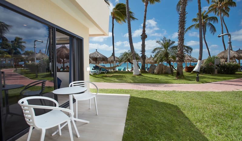 Beachside Room Queen Queen Interior of a modern hotel room featuring two blue velvet lounge chairs and a white coffee table, with a large window and sliding glass door overlooking a tropical beach with palm trees and hammocks at Divi Aruba All Inclusive Resort by Kenwood Travel