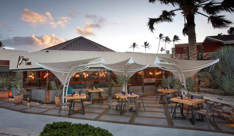 Pure Lime restaurant at night, featuring modern glass architecture, warm hanging lights, and outdoor patio seating under palm trees at Divi Aruba All Inclusive Resort by Kenwood Travel