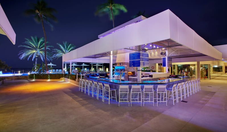 Pizza per Tutti outdoor beach bar at a luxury resort at night, featuring a sleek square counter with blue LED accent lighting, white bar stools, and palm trees under a dark sky in the background at Divi Aruba All Inclusive Resort by Kenwood Travel
