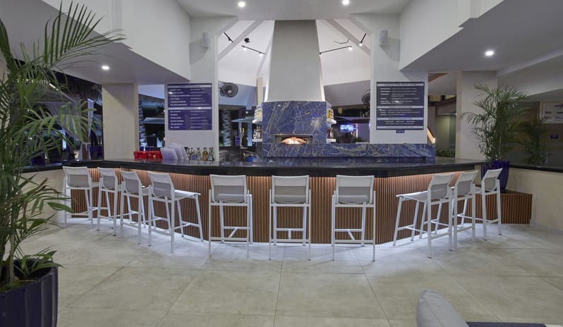 Coco Grill & Bar Modern resort bar with white high-back stools, wood-paneled counter, and a central blue marble pizza oven at Divi Aruba All Inclusive Resort by Kenwood Travel