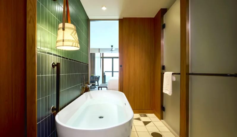 Mirage Premium Explorer (King) bathroom featuring a white freestanding soaking tub against a green-tiled wall with a woven pendant lamp, looking through to a bright bedroom and ocean-view balcony at Centara Grand Mirage Beah Resort by Kenwood Travel