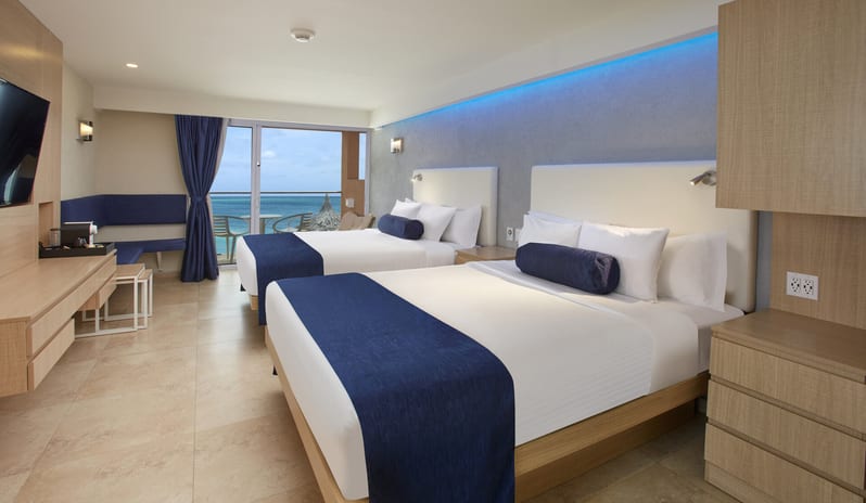 Deluxe Oceanfront Queen Queen beds, blue decorative pillows, and a private balcony overlooking the turquoise ocean at Tamarijn Aruba All Inclusive Resort by Kenwood Travel