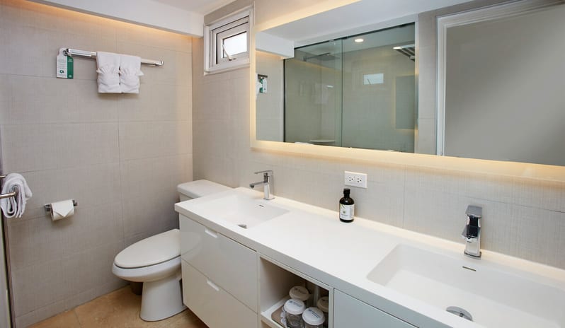 Deluxe Oceanfront Queen Queen A modern, clean hotel bathroom featuring a double white vanity, large backlit mirror, glass walk-in shower, and neutral tile walls at Tamarijn Aruba All Inclusive Resort by Kenwood Travel