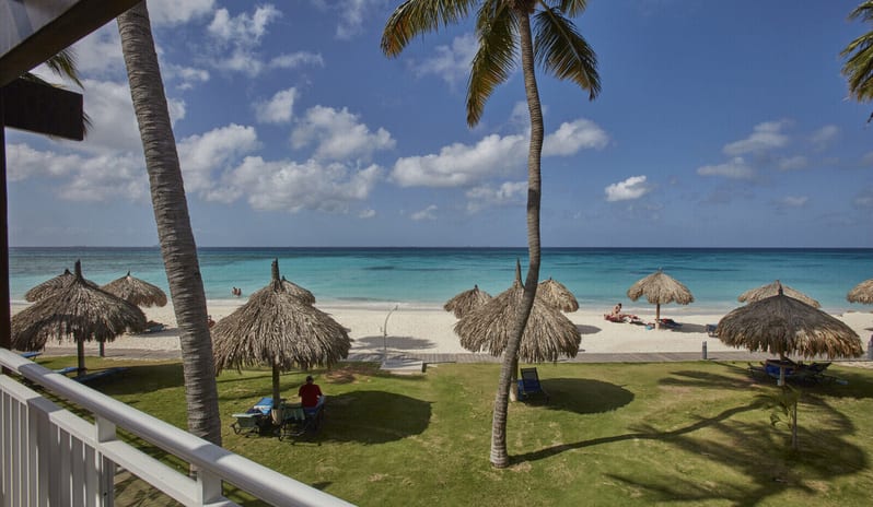 Deluxe Oceanfront Queen Queen hotel lounge with wicker papasan chairs and a blue sofa, opening through sliding glass doors to a private patio and white sand beach at  Tamarijn Aruba All Inclusive Resort by Kenwood Travel