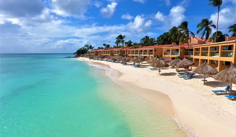 Deluxe Oceanfront Queen Queen An aerial perspective of a tropical beachfront resort with yellow buildings stretching along a white sand shoreline at Tamarijn Aruba All Inclusive Resort by Kenwood Travel