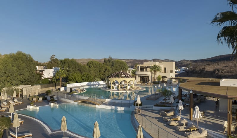 The Spiritzeria large turquoise swimming pool, a central domed gazebo bar, sun loungers, and scenic mountain views at Aulūs Lindos Rhodes, Curio Collection by Hilton by Kenwood Travel